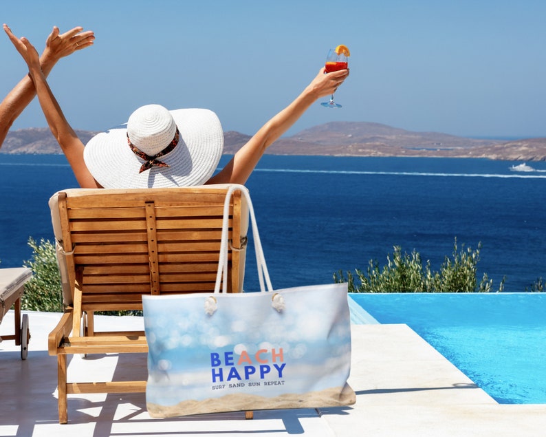May include: A woman relaxes in a lounge chair by a pool with a blue and white beach bag that says "Beach Happy Surf Sand Sun Repeat".