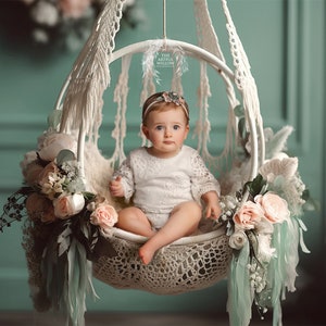 May include: A baby girl wearing a white lace dress sits in a white macrame swing decorated with pink and white flowers. The swing is hanging from the ceiling against a light green wall.
