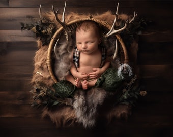 May include: A newborn baby boy is sleeping in a wicker basket with antlers and greenery. The baby is wearing a plaid shirt and a green and brown blanket. The basket is sitting on a brown fur rug.
