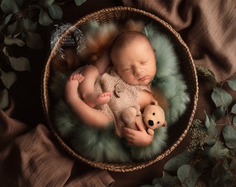 May include: A newborn baby sleeping in a woven basket with a plush green and brown fur lining. The baby is holding a small teddy bear.