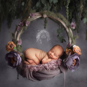 May include: A newborn baby sleeping peacefully in a purple and brown floral wreath, surrounded by greenery. The wreath is hanging from a branch.