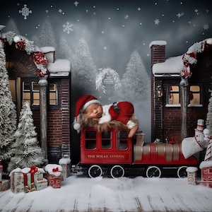 May include: A sleeping baby dressed as Santa Claus is on a red toy train with the words "The Polar Express" on the side. The train is in a snowy winter scene with two brick buildings and Christmas trees.