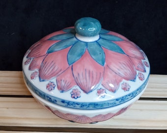 May include: A ceramic trinket box with a lid. The lid and base are white with a pink and blue floral design. The lid has a teal knob. The box is on a light wood surface.