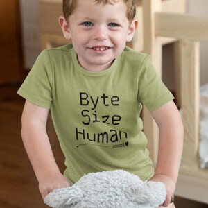 Op de afbeelding: Een jong kind draagt een lichtgroen t-shirt met de zwarte tekst "Byte Size Human". Het kind lacht en houdt een knuffel vast. Het shirt heeft de tekst "xoxox" onder het woord "Human".