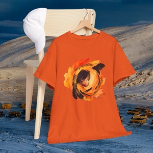 May include: Orange t-shirt with a watercolor illustration of a child's profile within a circular design. The design includes the text "Every child matters" and other phrases. A white baseball cap rests on a wooden chair.