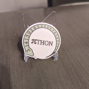 May include: A white circular sticker with a green snake wrapped around the edge. The snake's tail is at the bottom of the sticker and its head is at the top. The snake is holding a red and white 'pi' symbol in its mouth. The sticker has the text 'PYTHON' written in red letters inside the snake's body. The sticker is on a clear plastic stand.