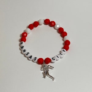 Könnte beinhalten: Rotes und weißes Perlenarmband mit den Worten "Kansas City" und einem silbernen Fußballspieler-Anhänger.
