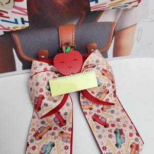 May include: A school-themed hair bow with a red apple and ruler embellishments. The bow features a light pink ribbon with a pattern of school buses, pencils, and flowers. The edges are trimmed with red ribbon.