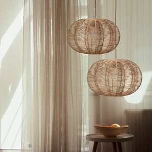 May include: Two woven rattan pendant lights hang from the ceiling. The spherical lampshades have an open weave design. A small, round side table with a wooden bowl and fruit sits below the lights. Sheer curtains and a window are in the background.