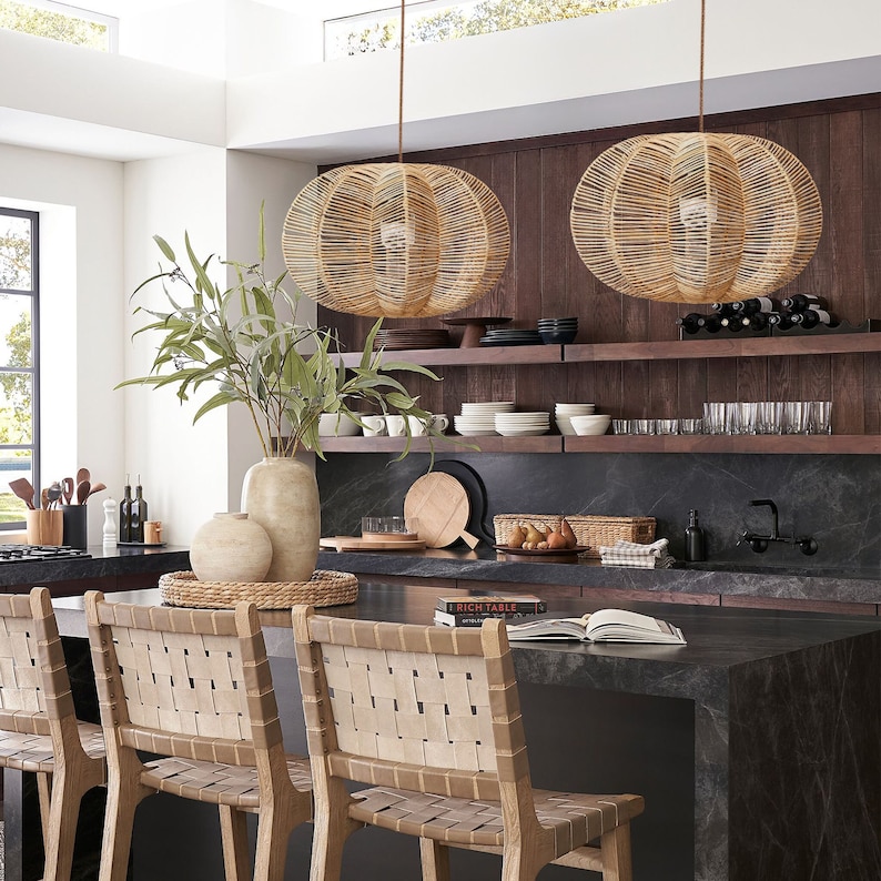 May include: Two woven pendant lights hang above a kitchen island with woven leather chairs. The kitchen features dark wood shelving with dishware, glassware, and a wine rack. A vase with greenery and decorative items sit on the island.
