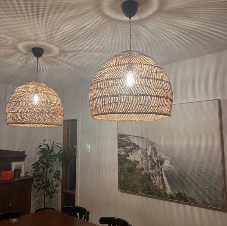 May include: Two woven wicker pendant lights hang from the ceiling over a dining table. The lights are brown and have a natural woven texture. The lights are on and casting a warm glow.