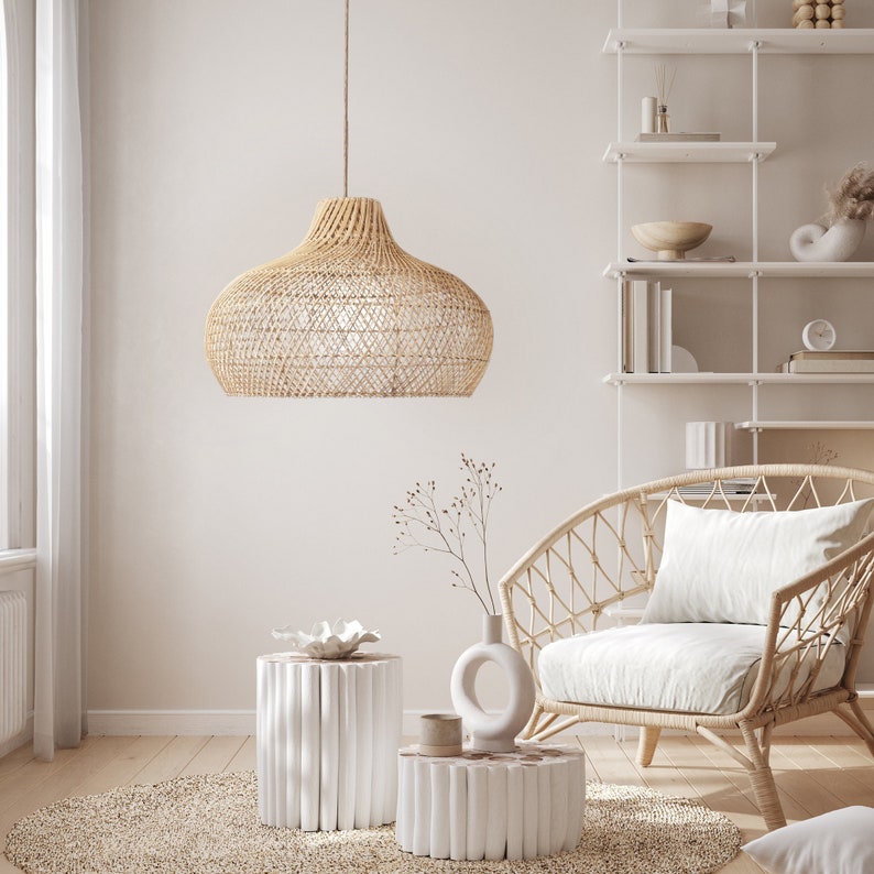 May include: A woven wicker pendant light hangs over a white wicker chair with a white cushion. Two white side tables with a woven design are in the foreground. The room is decorated in a minimalist style with a white wall and a white shelf unit.