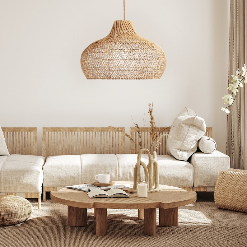 May include: A light brown wicker pendant lamp hangs over a white sofa with wooden slats. The sofa has a white throw pillow and a brown throw pillow. A wooden coffee table with a woven top sits in front of the sofa. The table has a white ceramic candle, a small white cup, and a book on top.