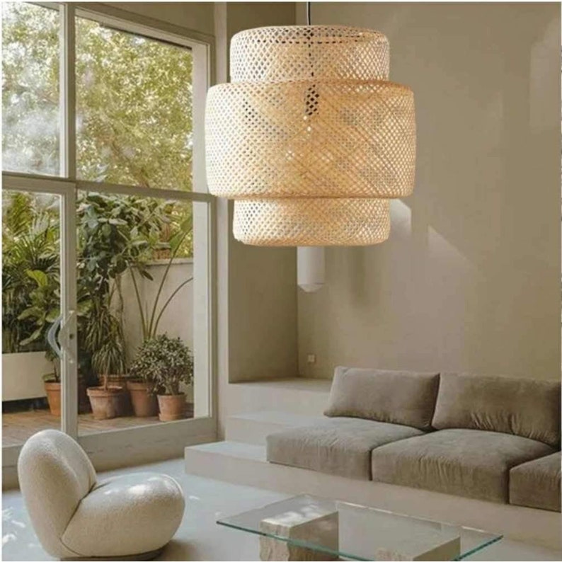 May include: A three-tiered woven bamboo pendant light fixture hangs from the ceiling in a modern living room. The light fixture is made of natural woven bamboo and has a warm, natural color. The light fixture is hanging over a light brown couch with a glass coffee table in front of it.