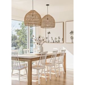May include: A dining room with a wooden table and white chairs. Two large, woven, natural-toned pendant lights hang above the table. The room has a coastal theme with artwork featuring palm trees.