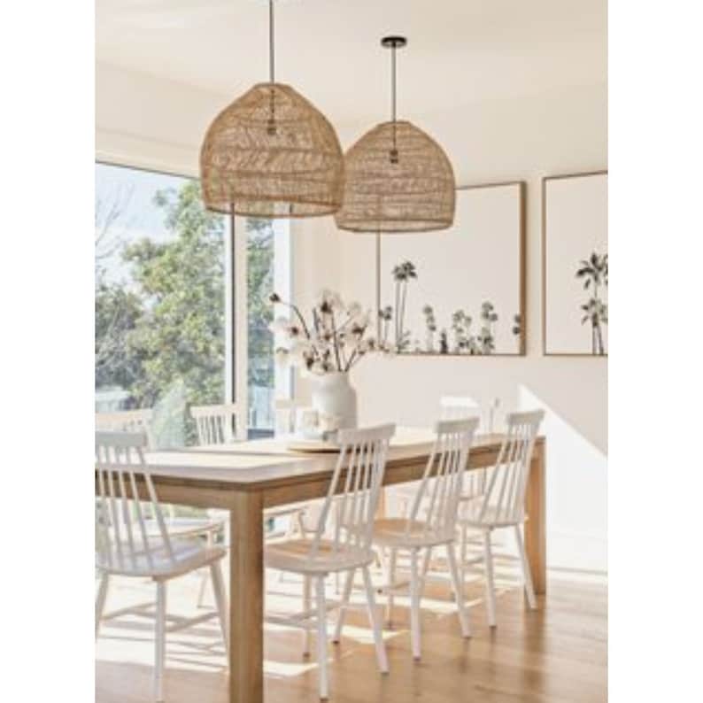 May include: A dining room with a wooden table and white chairs. Two large, woven, natural-toned pendant lights hang above the table. The room has a coastal theme with artwork featuring palm trees.