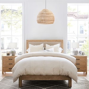 May include: A light wood bed frame with a white linen duvet and two white pillows. The bed is in a bedroom with two windows and a wicker pendant light hanging above the bed.