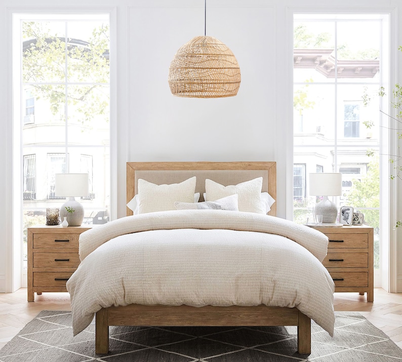 May include: A light wood bed frame with a white linen duvet and two white pillows. The bed is in a bedroom with two windows and a wicker pendant light hanging above the bed.
