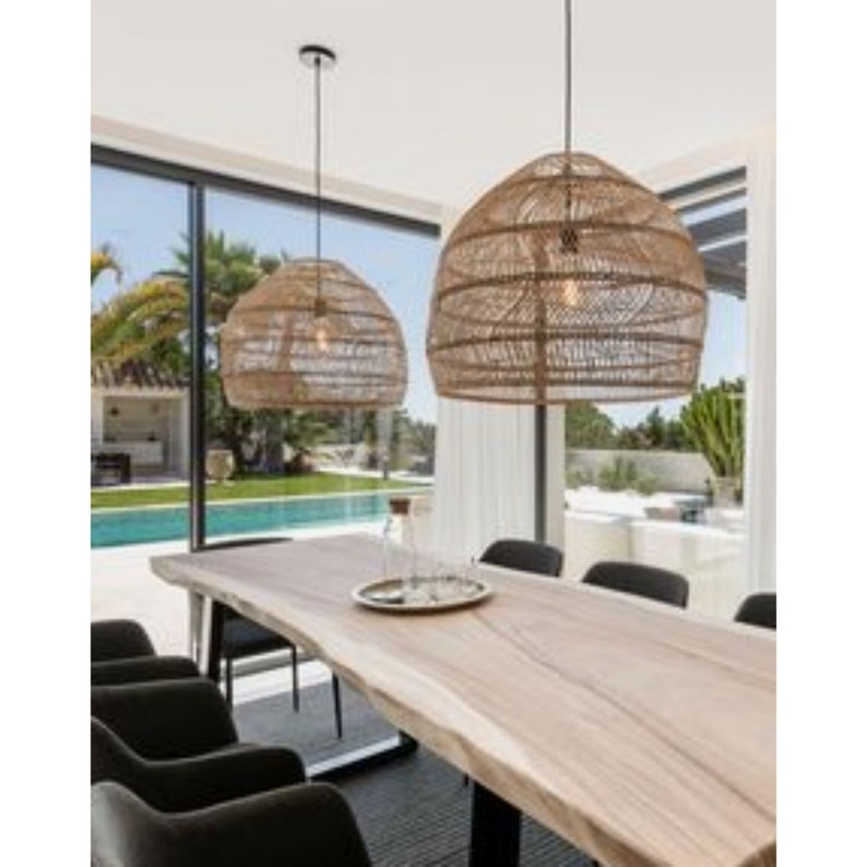 May include: Two woven rattan pendant lights hang over a long wooden table with black chairs in a modern dining room. The table has a live edge design and the chairs are upholstered in a dark gray fabric.