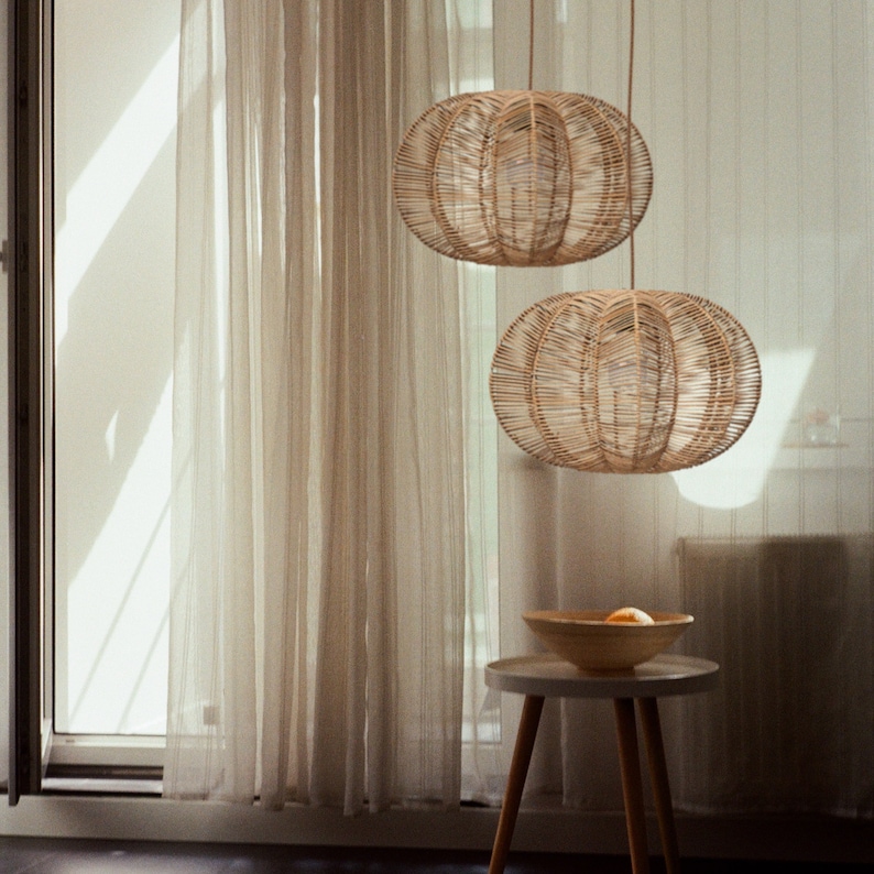 May include: Two woven rattan pendant lights hang from the ceiling. The spherical lampshades have an open weave design. A small, round side table with a wooden bowl and fruit sits below the lights. Sheer curtains and a window are in the background.