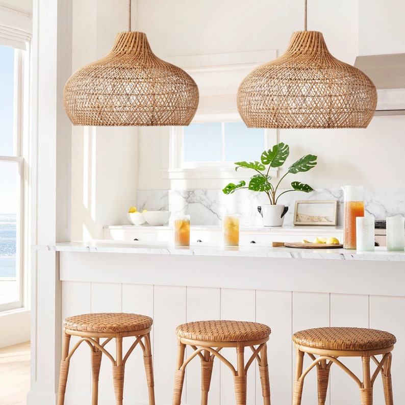 May include: Two woven rattan pendant lights hang over a kitchen island with three woven rattan bar stools. The island has a white countertop and white cabinets.