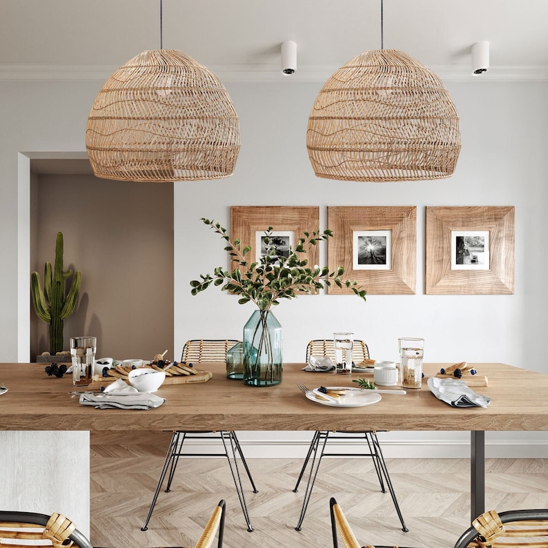 May include: Two woven wicker pendant lights hang over a wooden dining table with six chairs. The table is set with plates, glasses, and a vase of greenery.