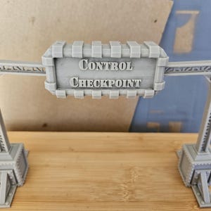 Könnte beinhalten: Ein graues 3D-gedrucktes Modell eines Kontrollstellen-Schildes. Das Schild trägt die Aufschrift "CONTROL CHECKPOINT" in Großbuchstaben. Das Modell hat zwei Stützstrukturen auf beiden Seiten des Schildes mit detailliertem Design. Das Modell steht auf einer Holzoberfläche.