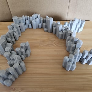 May include: A grey, 3D-printed terrain piece resembling a rocky mountain range. The piece is composed of numerous hexagonal and rectangular shapes, creating a jagged, textured surface. The terrain is displayed on a wooden surface.