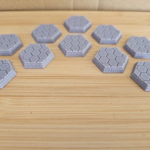 May include: A set of twelve hexagonal game pieces made of a light purple plastic. The pieces are arranged in a semi-circle on a wooden surface.