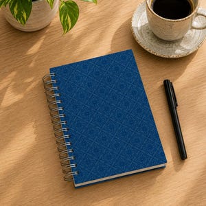 May include: A spiral-bound notebook with a deep blue cover featuring a repeating geometric floral pattern. The notebook is positioned alongside a black ballpoint pen, a ceramic cup filled with black coffee, and a small potted green plant.