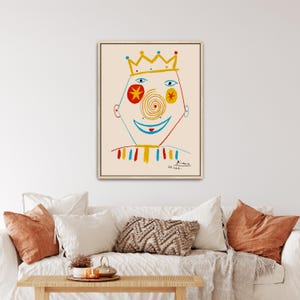 May include: A framed wall art print featuring a stylized line drawing of a face wearing a yellow crown. The face has a red star-shaped eye, a blue eye, a spiral nose, and a blue smile. The figure wears a striped collar in red, yellow, and blue.