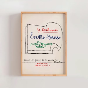 May include: A framed art print featuring the text "Le Corbusier Entre-deux" in red, blue, and green. The artwork has a minimalist style with handwritten text and a simple black outline. The frame is made of light-colored wood.