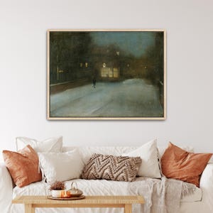 May include: Framed art print of a nighttime winter scene. A lone figure walks down a snow-covered street, illuminated by the warm glow of building lights. The artwork is framed in a light-colored wooden frame.