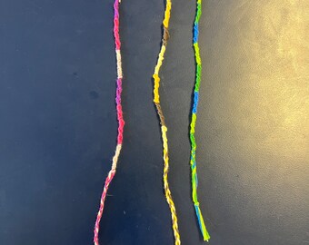 May include: Three handmade friendship bracelets in vibrant colors. One is pink, white, and purple. Another is brown, yellow, and tan. The third is green, blue, and yellow. Each bracelet is made of braided thread.