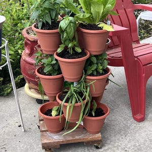 Puede incluir: Una jardinera escalonada de color terracota llena de varias hierbas y verduras de hoja verde. Las macetas apiladas se asientan sobre un peque&ntilde;o palet de madera con ruedas, creando un jard&iacute;n vertical. Una silla de pl&aacute;stico roja est&aacute; en el fondo.