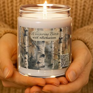 Puede incluir: Una vela blanca encendida en un tarro de cristal. La etiqueta dice "Whispering Birch with Affirmations" y presenta un diseño de abedul. La vela se sostiene en manos ahuecadas.