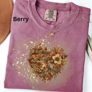 以下が含まれることがあります： ベリー色のTシャツ、ハート型の花柄デザイン。茶色、ベージュ、緑色の様々な花、葉、ドングリがデザインされ、金色のグリッターアクセントが施されています。ペンダント付きのシルバーネックレスがシャツの上にあります。