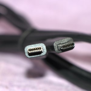 Könnte beinhalten: Nahaufnahme eines schwarzen USB-Kabels mit zwei verschiedenen Steckertypen. Die Stecker haben mehrere Pins für die Datenübertragung. Das Kabel ist aufgerollt und scheint aus flexiblem Material zu bestehen. Der Hintergrund ist ein weiches, unscharfes Rosa.