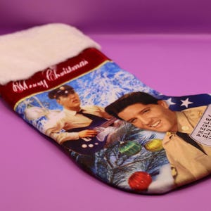 May include: Christmas stocking with images of Elvis Presley. The stocking has a white faux fur cuff, a red top with "Merry Christmas" text, and gold tassels. The design includes a star and stripes.