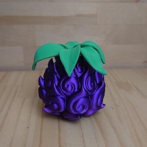 May include: A purple, stylized fruit with a textured surface and green leaves. The fruit has a unique, swirling pattern and is set against a light wooden background. The design is reminiscent of a fantasy or anime theme.