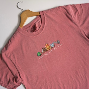 May include: A pink t-shirt with a pixelated design of a video game character and the text "fighting for my life".