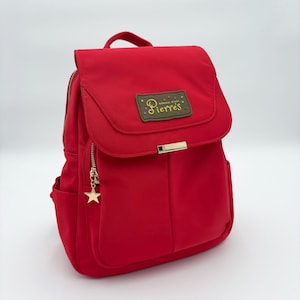 Puede incluir: Una mochila roja brillante con cierre de solapa y un asa pequeña. La mochila tiene una cremallera dorada y un charm en forma de estrella. La solapa tiene el texto "Pierres General Store".