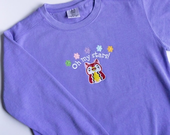 Puede incluir: Sudadera morada con un búho bordado de colores y el texto "Oh my stars!"