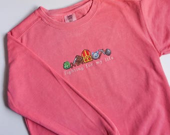Puede incluir: Una sudadera rosa con un diseño bordado en blanco que presenta una fila de iconos de videojuegos pixelados y el texto "fighting for my life".