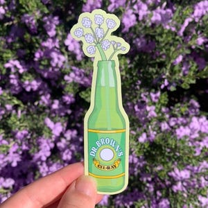 May include: A sticker featuring a green bottle of Dr. Brown's Cel-Ray soda with a bouquet of white flowers. The sticker has a yellow border and is held against a backdrop of purple flowers.