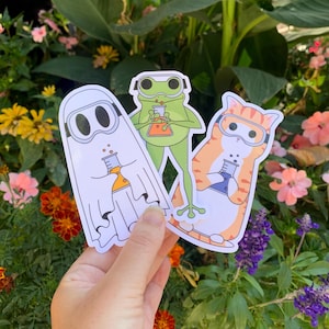 May include: Three colorful stickers featuring a ghost, a frog, and a cat, each wearing goggles and holding a beaker. The stickers are on a white background and have a cartoon style.