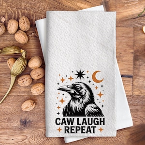 May include: White kitchen towel with a black raven illustration, stars, and a crescent moon. The text "CAW LAUGH + REPEAT +" is below the raven. The towel is folded on a wooden surface with walnuts and nutcrackers.