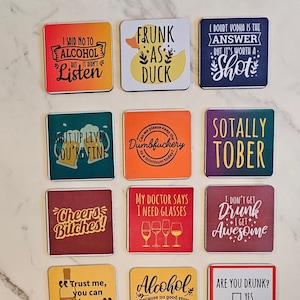 May include: Ten colorful square coasters with humorous sayings about alcohol. The coasters feature various designs, including a duck, glasses of wine, and a check box with the text "Are you drunk?"