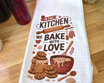 Può includere: Canovaccio da cucina bianco con la scritta "IN THIS KITCHEN WE BAKE WITH LOVE". Il canovaccio presenta illustrazioni di utensili da forno, biscotti e una ciotola. Il canovaccio è piegato e posto su una superficie di legno.