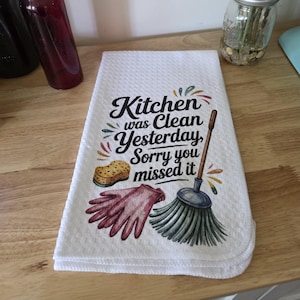 May include: White kitchen towel with the text "Kitchen was Clean Yesterday, Sorry you missed it." The towel features a sponge, a pink glove, and a broom graphic. The towel is folded on a wooden surface.